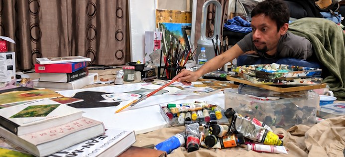 Davey Jose lying on the studio floor, reaching out with a brush to paint a canvas surrounded by oil paints and art books