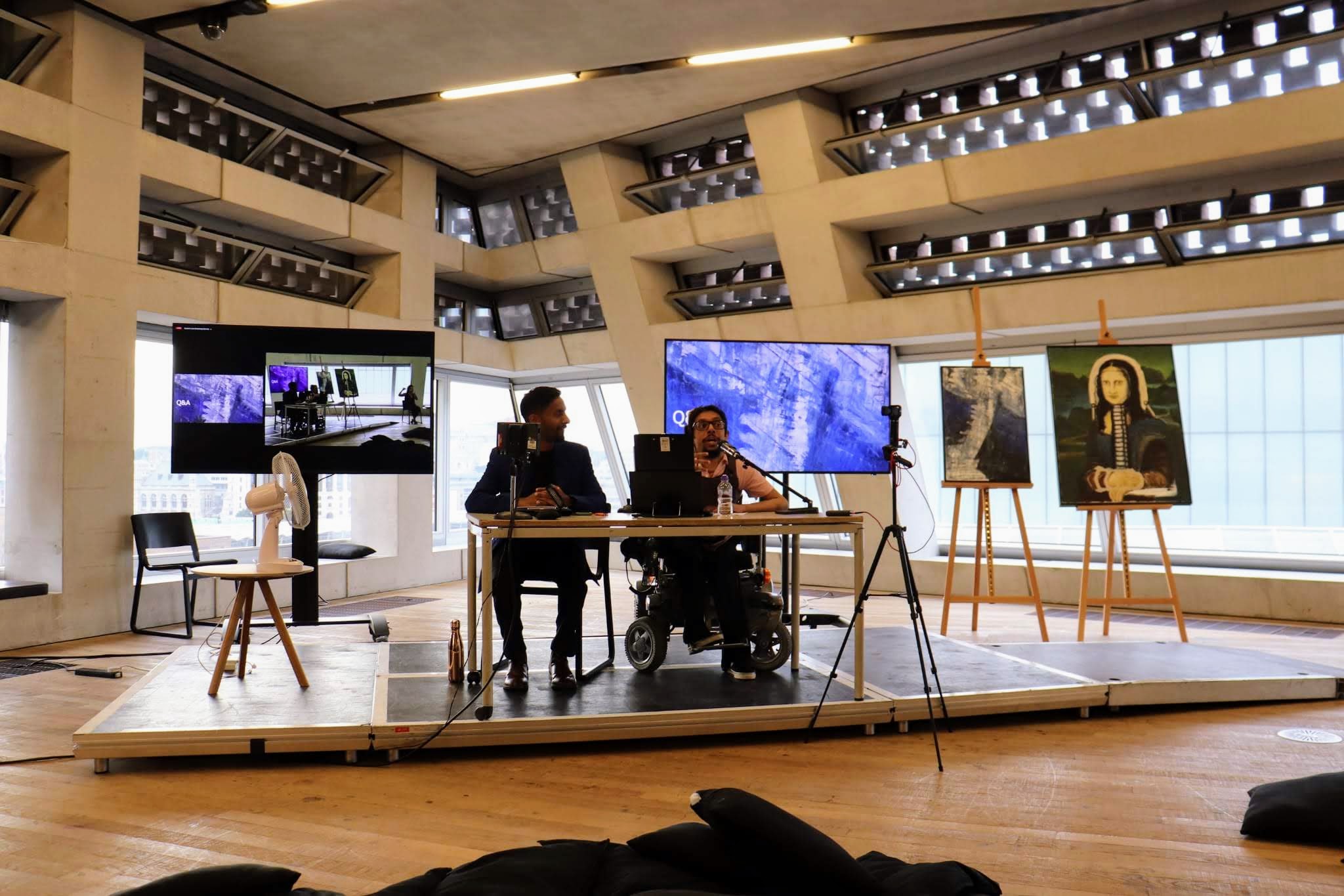 Davey Jose and Bobby Seagull on stage at Tate Modern, with oil paintings on easels and the work projected on screens behind them
